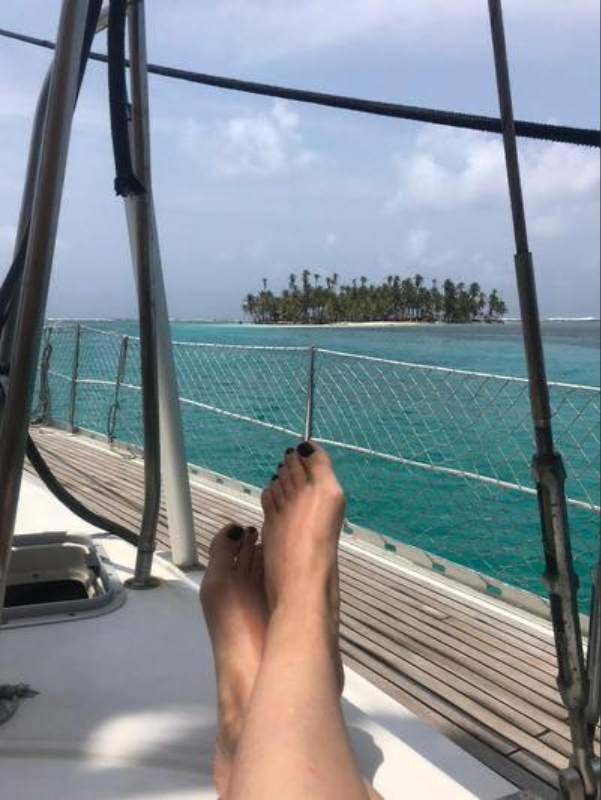A bordo de un velero en Guna Yala, Panamá Pies de una persona descansando en la cubierta de un velero, frente a una pequeña isla tropical rodeada de aguas turquesas en Guna Yala, Panamá.