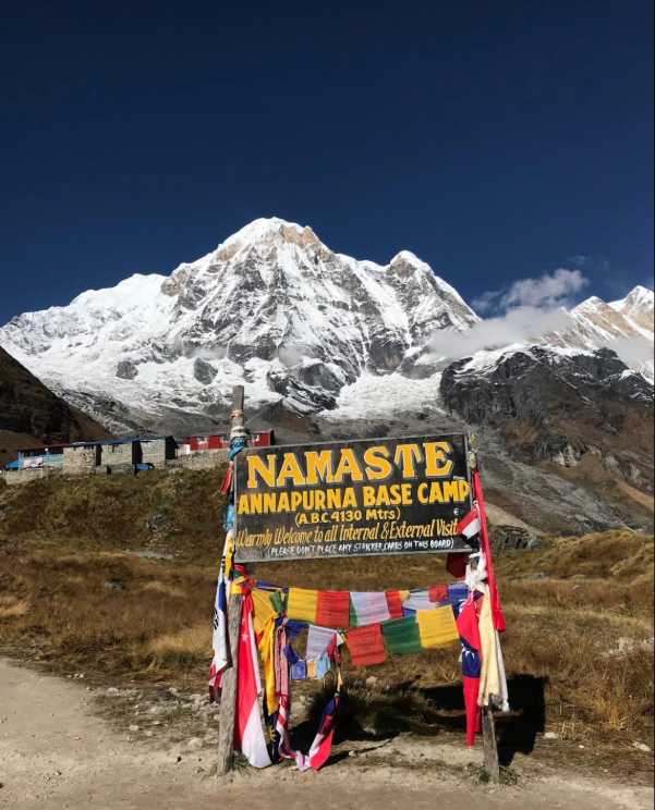 Campamento Base del Annapurna, Nepal: un paisaje del Himalaya Cartel del Campamento Base del Annapurna a 4.130 metros, decorado con banderas de oración, frente a una imponente montaña del Himalaya cubierta de nieve.