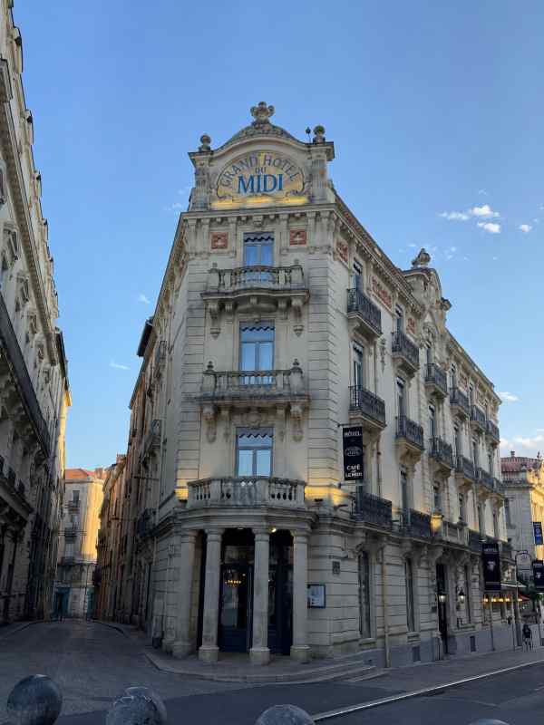 Fotografía del Grand Hôtel du Midi, en el centro de Montpellier, Francia. El edificio destaca por su elegante fachada de estilo histórico, ricamente ornamentada, con numerosos balcones, columnas y detalles decorativos. En la parte superior puede leerse el nombre del hotel, coronado por elementos escultóricos. La fachada se encuentra en la esquina de una calle, bajo un cielo azul y despejado, rodeada por otros edificios históricos del centro de la ciudad.