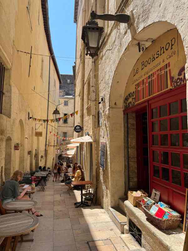 Una estrecha calle del casco histórico de Montpellier, Francia, flanqueada por antiguos edificios de piedra. En primer plano destaca Le Bookshop, una librería anglófona y café con una llamativa fachada de color rojo y libros expuestos en cestas junto a la entrada. La calle está decorada con banderines de colores y faroles tradicionales, mientras varias terrazas de cafés se extienden a lo largo del callejón. La luz del sol y la arquitectura de piedra crean una atmósfera cálida y acogedora.