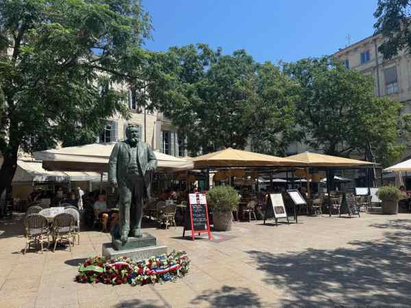 Place Jean Jaurès, el corazón de Montpellier La Place Jean Jaurès es una animada plaza del centro de Montpellier, Francia. En el centro de la imagen aparece una estatua de Jean Jaurès, rodeada de flores depositadas a sus pies. Alrededor de la plaza se encuentran varias terrazas de cafés y restaurantes, protegidas por grandes sombrillas. Los árboles proporcionan sombra a los visitantes y los edificios históricos que rodean la plaza completan el ambiente urbano y mediterráneo de este popular punto de encuentro de Montpellier.