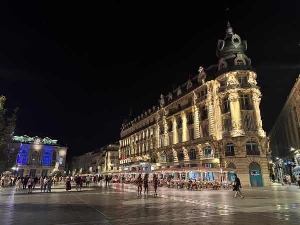 Vista nocturna de la Place de la Comédie, en el centro de Montpellier, Francia. La amplia plaza está iluminada y rodeada de edificios históricos con fachadas ornamentadas. Destaca especialmente un imponente edificio de estilo historicista, profusamente decorado y con una torre en la esquina. A su alrededor hay terrazas de cafés y restaurantes llenas de visitantes, mientras numerosas personas pasean por la gran explanada. Al fondo se observan otros edificios iluminados que completan la animada escena nocturna.