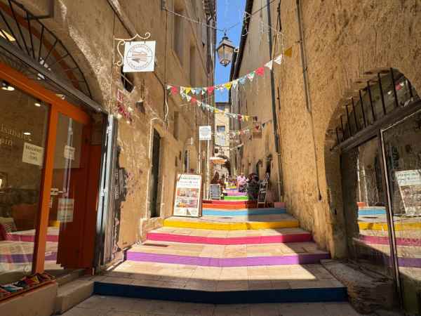 La Rue du Bras-de-Fer, en el centro histórico de Montpellier, es una de las calles más singulares y fotografiadas de la ciudad. La estrecha calle medieval está flanqueada por edificios de piedra y decorada con una llamativa escalinata pintada en franjas de colores rosa, amarillo, azul, naranja y morado. Sobre la calle cuelgan pequeñas banderolas de colores y antiguos faroles, mientras tiendas y restaurantes ocupan los bajos de los edificios.
