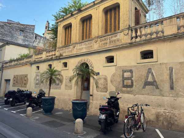 Fotografía de un antiguo edificio de los baños públicos de Montpellier, Francia, que conserva en su fachada las inscripciones relacionadas con los antiguos “Bains”. El edificio, de color ocre y con una arquitectura sencilla de inspiración mediterránea, presenta grandes ventanales con persianas de madera y una terraza con balaustrada. En la planta baja se encuentra actualmente el restaurante Pampa. Frente al edificio hay varias motocicletas, bicicletas y grandes maceteros con palmeras.