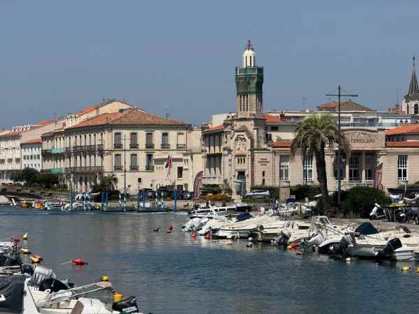 Vista del canal portuario de Sète, en el sur de Francia, flanqueado por embarcaciones de recreo amarradas en fila a ambos márgenes. En la orilla se alzan edificios históricos de fachada clara con balcones de hierro forjado y tejados de teja roja, junto a un edificio institucional con una torre-reloj coronada por una cúpula verde. Una palmera y algunas banderas completan la escena, bajo un cielo azul despejado propio de un día soleado de verano.