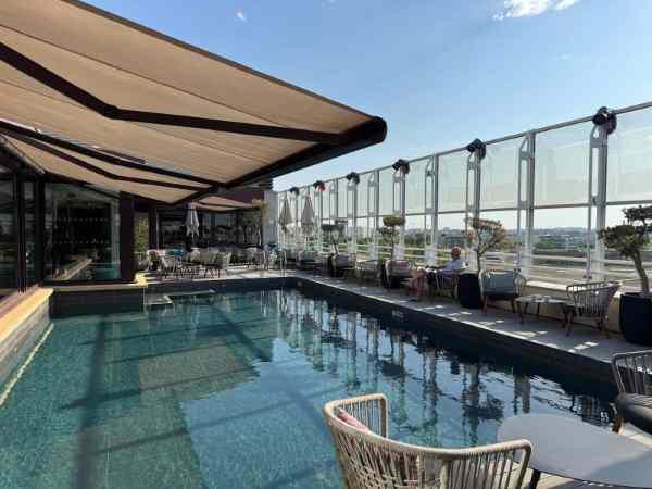 Piscina situada en la azotea del Hotel Pullman de Montpellier, protegida por un amplio toldo retráctil de rayas. Alrededor del vaso se distribuyen sillones y mesas de exterior sobre un suelo de tarima, junto a un cerramiento acristalado que ofrece vistas panorámicas de la ciudad. La luz cálida y el cielo despejado dan al espacio un ambiente relajado, propio de una terraza de hotel en verano.