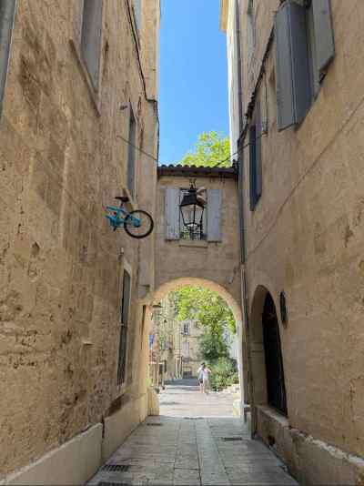 Vista de una callejuela típica del centro histórico de Montpellier, con altas fachadas de piedra caliza y contraventanas grises. Una bicicleta azul cuelga decorativamente de la pared izquierda, junto a un farol de hierro forjado. Al fondo, un arco de piedra da paso a otra calle donde se distingue una persona caminando y vegetación verde bajo un cielo despejado.