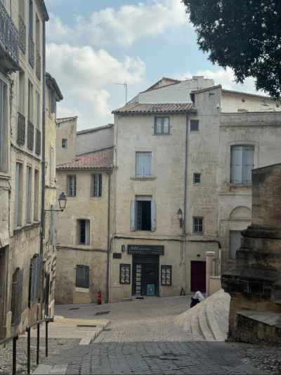 Vista de una pequeña plaza en el casco histórico de Montpellier, con edificios de piedra caliza de varias alturas y contraventanas azules. En primer plano, unas escaleras de piedra descienden hacia una calle empedrada donde se aprecia una farola clásica y un hidrante rojo. El cielo parcialmente nublado y un árbol frondoso enmarcan la escena.