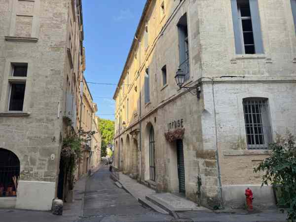 Fotografía de una calle estrecha y empinada en el barrio histórico de l'Écusson, en Montpellier, Francia. A ambos lados se alzan edificios de piedra caliza con contraventanas de madera, balcones de hierro forjado y una farola clásica. En el edificio de la derecha destaca el letrero "INFUSE" sobre la fachada. Al fondo se ve el cielo azul despejado, árboles verdes y algunos transeúntes caminando por la calle.