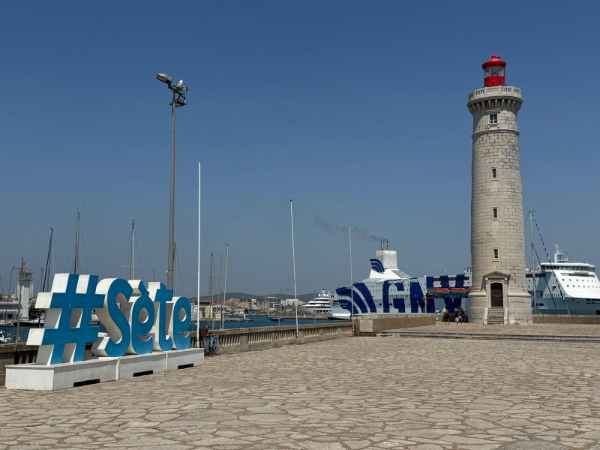 Fotografía del puerto de Sète, en el sur de Francia. En primer plano aparece un gran cartel con el nombre de la ciudad, “Sète”, junto al paseo marítimo. Destaca un alto faro de piedra coronado por una linterna roja. Al fondo se observan veleros, embarcaciones y grandes barcos atracados en el puerto, bajo un cielo azul y despejado.