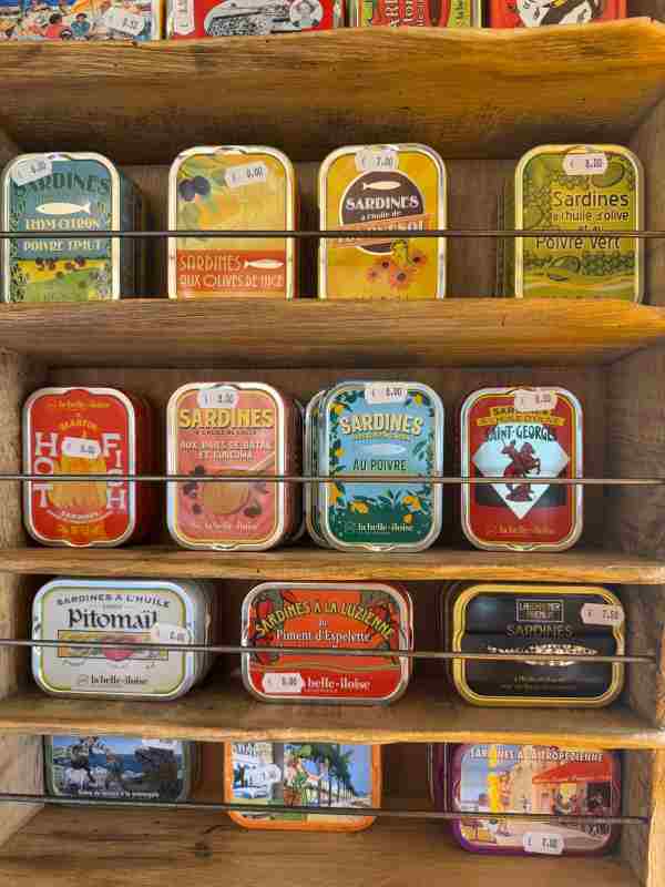 Fotografía de una estantería de madera en una tienda de productos gourmet en Sète, Francia, con múltiples filas de latas de sardinas de la marca La Belle-Iloise. Las latas presentan diseños coloridos e ilustrados con distintos sabores, como sardinas al tomillo y limón, a las aceitunas de Niza, al pimentón de Espelette, con miel de flores de Provenza y sardinas picantes "Hot Fish", entre otras. Cada lata muestra su precio en una etiqueta blanca, con valores que oscilan entre 7,00 € y 9,50 €.