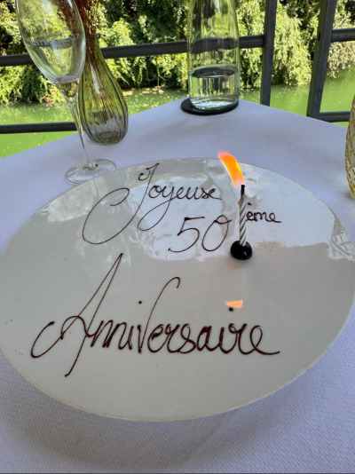 Plato de postre decorado con la frase "Joyeuse 50ème Anniversaire" (Feliz 50 aniversario) escrita con chocolate en letra cursiva, junto a una vela encendida en su base negra. Al fondo, sobre la mesa cubierta con mantel blanco, se ven una copa de champán, un jarrón con flores secas y una botella, con un jardín verde y una barandilla de hierro de fondo.