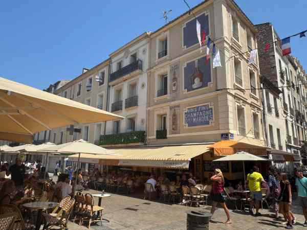 Fotografía tomada en el mercado de Sète, en el sur de Francia. La imagen muestra una animada plaza peatonal rodeada de cafés, bares y restaurantes con numerosas terrazas y sombrillas. Los edificios de varias plantas presentan fachadas tradicionales y carteles decorativos relacionados con el vino y el champán. En las calles hay vecinos y visitantes disfrutando del ambiente soleado de esta localidad mediterránea.