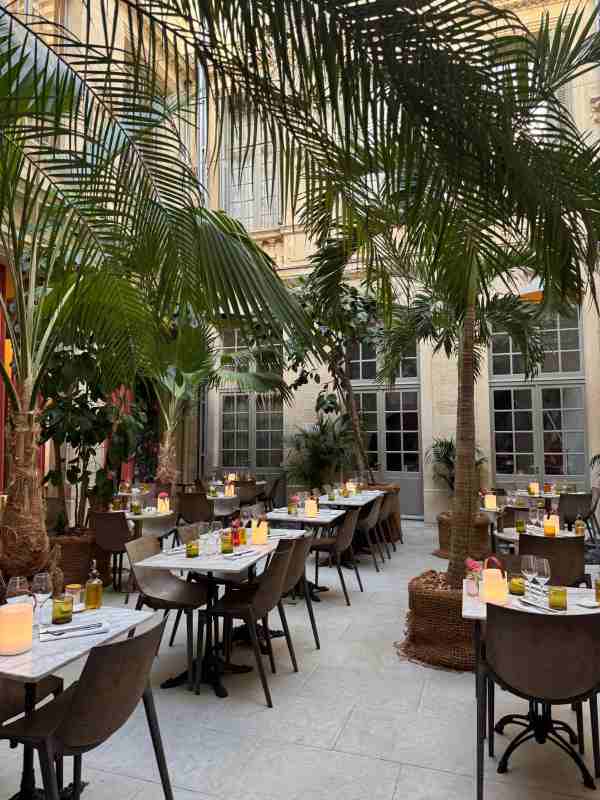 Fotografía del patio interior del restaurante del Hôtel Richer de Belleval, en Montpellier. El espacio está decorado con varias mesas de mármol redondas y rectangulares, sillas de diseño en tonos marrón oscuro, velas encendidas dentro de vasos y pequeños ramos de flores. Grandes palmeras y plantas tropicales cubren el techo del patio, creando un ambiente fresco y sombreado. Al fondo se aprecia la fachada de piedra del hotel con puertas y ventanas de estilo francés en tonos grises.
