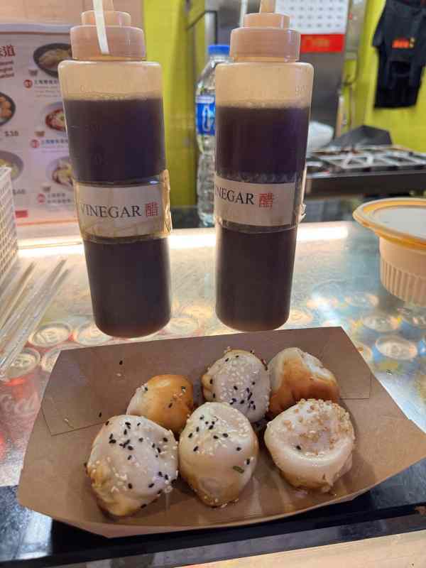 Seis sheng jian bao, panecillos chinos rellenos y dorados a la plancha, servidos en una bandeja de cartón con semillas de sésamo y dos botellas de vinagre al fondo. Copiar ALT