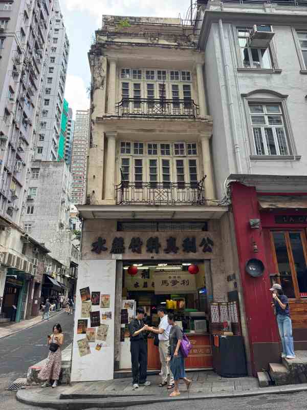 Antiguo edificio comercial de Hong Kong con fachada envejecida, balcones de hierro forjado y una tienda con letreros chinos en la planta baja, junto a una calle en pendiente.