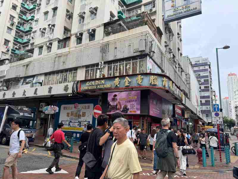 Fachada del Golden Computer Arcade, en Sham Shui Po, Hong Kong, con grandes letreros en chino e inglés, edificios residenciales y numerosos peatones cruzando la calle.