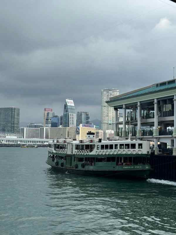 Ferry verde y blanco de la compañía Star Ferry junto al muelle de Hong Kong, con los rascacielos de Kowloon al fondo y un cielo cubierto de nubes grises.