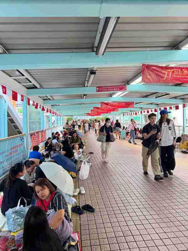 Pasarela peatonal cubierta de Hong Kong, con numerosas personas sentadas en el suelo y otras caminando bajo banderas chinas y hongkonesas.